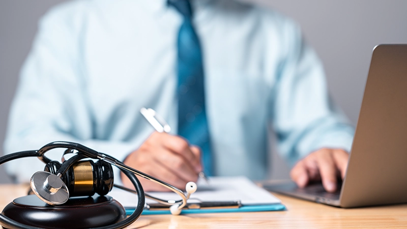 A lawyer or legal professional, is seated at a desk with a stethoscope and a judge's gavel draped and actively writing on a document on a clipboard with one hand while using a laptop with the other.