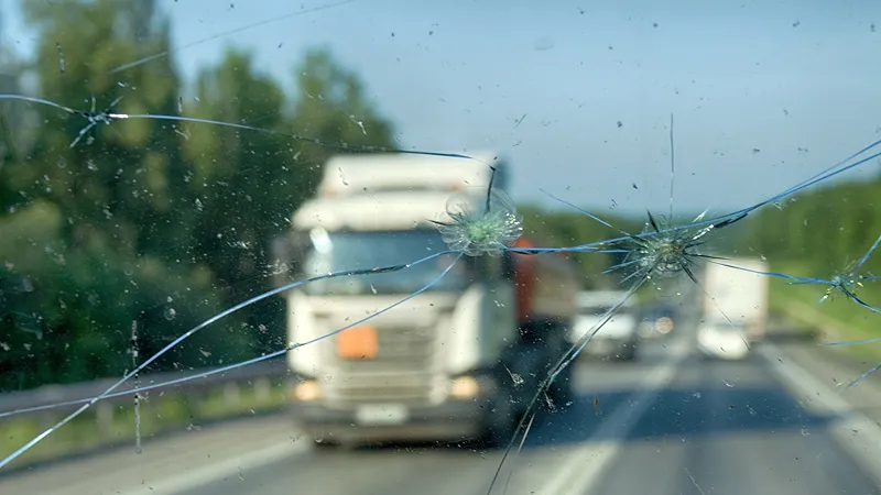 A large white semi-truck or heavy goods vehicle is on the road with a cracked and damaged windshield.